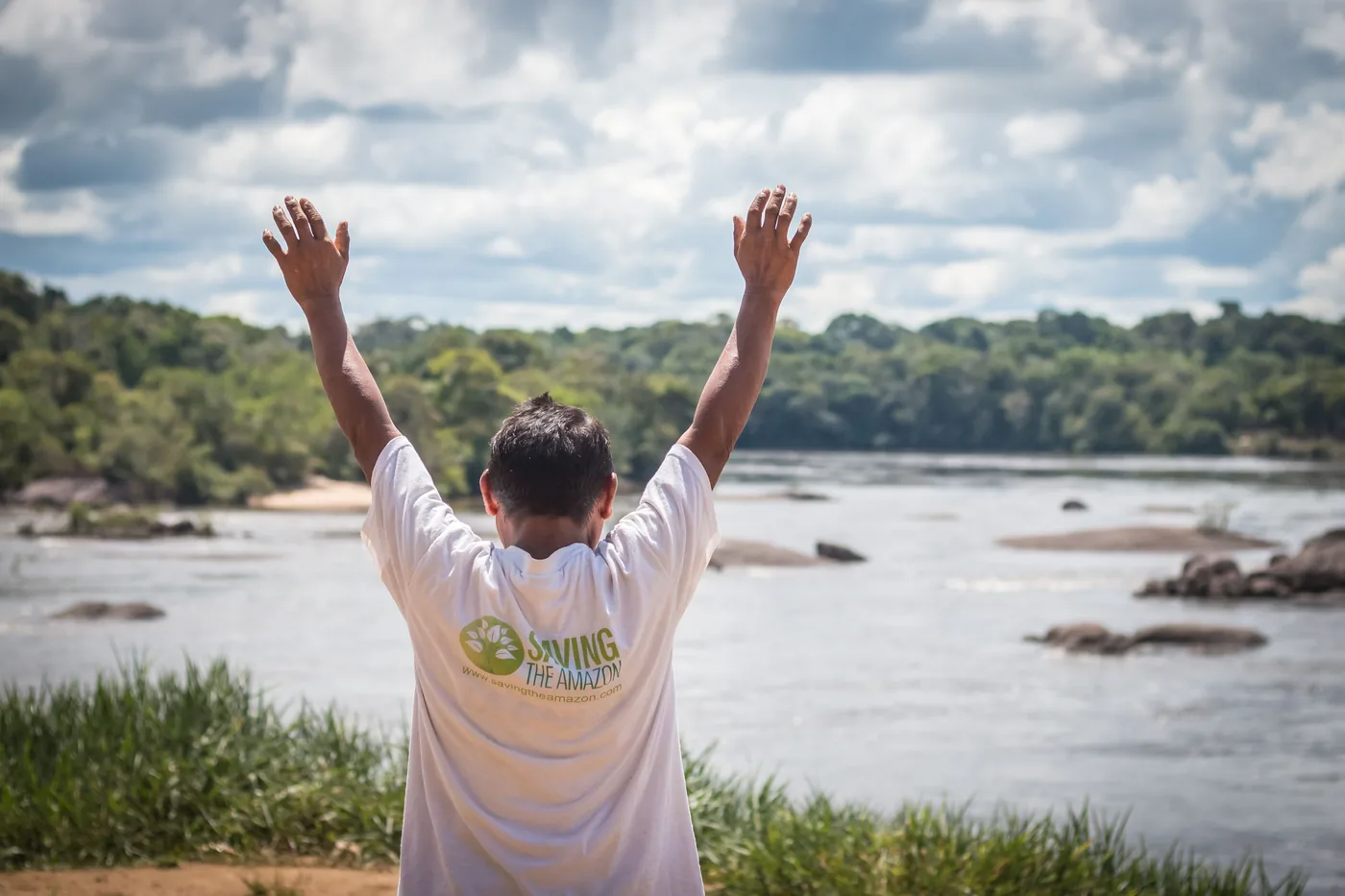 Integrante de uma comunidade parceira da Saving The Amazon com os braços erguidos diante de um rio, em um dos territórios de restauração